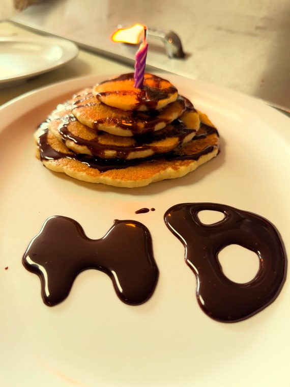 A stack of pancakes with a birthday candle and "HBD" written in chocolate sauce on the plate