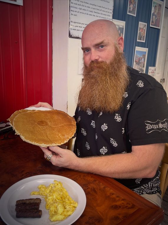 A bearded guest holds up a pancake bigger than his plate